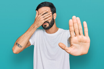 Wall Mural - Young hispanic man wearing casual white t shirt covering eyes with hands and doing stop gesture with sad and fear expression. embarrassed and negative concept.