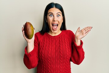 Beautiful brunette woman holding mango celebrating achievement with happy smile and winner expression with raised hand