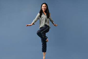 Wall Mural - Happy brunette girl in black and white striped shirt and wide trousers smiling and jumping on blue isolated background..