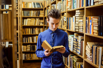 Thin guy in blue shirt reading a book with interest. Student in the library, university, college education, intelligent person.