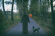 Mysterious man in monk or wizard costume holding orange air balloon with Jack-o-lantern pumpkin head image standing alone on autumn road. Halloween concept.