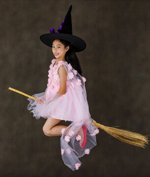 Portrait studio shot of little cute Asian kid in pink witch dress costume with black high hat look at camera posing flying gesture riding magic witchcraft broomstick on Halloween traditional festival
