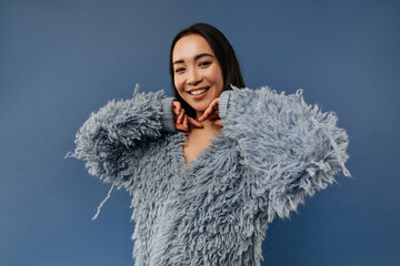 Wall Mural - Charming asian lady with cute smile and tanned skin in fashionable fluffy sweater with long sleeve looking into camera on blue backdrop..