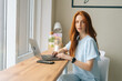 Portrait of serious attractive young female freelancer typing text on keyboard using laptop sitting at table by window in cozy cafe. Pretty redhead Caucasian lady remote working or studying.