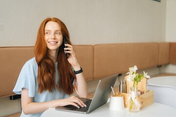 Portrtait of happy young woman talking on smartphone and using laptop sitting at table by window in cozy light cafe, looking away. Pretty redhead Caucasian lady remote working or studying.