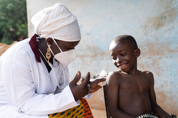 Black nurse with face mask injecting a dose of vaccine to a smiling brave schoolboy in an African hospital setting