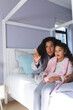 © Wavebreak Media - Happy mixed race mother and daughter sitting on bed, embracing and looking through window