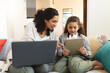 © Wavebreak Media - Smiling mixed race mother and daughter sitting on sofa, using laptop and tablet