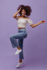 Canvas Print - Cheerful curly brunette woman in denim pants and white top dances on purple background. Attractive dark-skinned girl smiles.