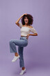 Good-humored dark-skinned woman in denim pants and cropped white top smiles widely, rises leg and poses on purple background.
