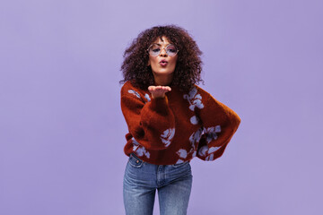 Poster - Pretty brunette woman in jeans, eyeglasses and burgundy sweater blows kiss on isolated. Charming lady poses on purple background.