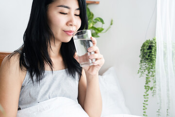 thirsty Asian woman drinking fresh water from glass after wakeup in morning sitting in bed