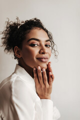 Poster - Beautiful dark-skinned lady in white blouse looks into camera. Portrait of curly brunette woman smiling in isolated grey background.