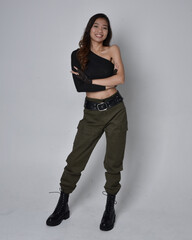 Full length portrait of pretty brunette, asian girl wearing black top and khaki utilitarian army pants and leather boots. Standing pose with gestural hands, isolated against a light grey studio backgr