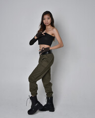 Full length portrait of pretty brunette, asian girl wearing black top and khaki utilitarian army pants and leather boots. Standing pose with gestural hands, isolated against a light grey studio backgr