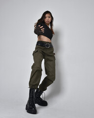 Full length portrait of pretty brunette, asian girl wearing black top and khaki utilitarian army pants and leather boots. Standing pose with gestural hands, isolated against a light grey studio backgr