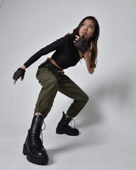 Full length portrait of pretty brunette, asian girl wearing black top and khaki utilitarian army pants and leather boots. Standing pose with gestural hands, isolated against a light grey studio backgr