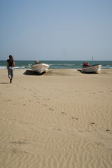 A woman silhouette seen from the back walking on a sand beach on a sunny windy day in a fishing village, two white boats on the beach and footsteps in sand