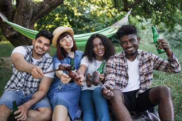 Wall Mural - Four joyful friends enjoying grilled sausages and cold beer during picnic on fresh air. Multiracial young people chilling together outdoors. Relaxation time.