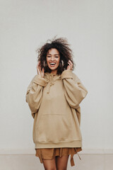 Happy dark-skinned girl smiles widely and listens to music in headphones on grey background. Curly woman in hoodie poses.