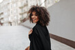 © Look! - Attractive African woman in black coat walks outside. Dark-skinned curly lady looks into camera and smiles outdoors.