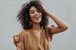 © Look! - Portrait of curly brunette African dark-skinned woman in beige top smiling and ruffling hair on isolated grey background.