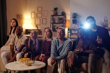 Wall Mural - Group of young intercultural friends sitting on long couch and watching movie