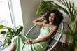 © Look! - Dark-skinned woman in green dress sits on chair. Curly brunette lady in silk outfit relaxes in armchair at home.
