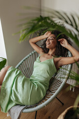 Poster - Cute brunette woman in silk dress smiles sincerely. Dark-skinned lady in green outfit relaxes and poses on armchair near white wall at home.