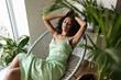 © Look! - Relaxed woman in silk dress sits on comfortable armchair. Young dark-skinned lady in green outfit poses in white room with plants.