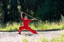 Beautiful Teen Girl In Red Sports Wushu Uniform Is Training Kung Fu In The Park,   Fighter Junior Athlete, Practicing Martial Arts With A Stick