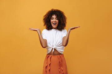 Wall Mural - Positive girl in summer outfit smiling on isolated backdrop. Woman in white t-shirt and orange skirt pointing to place for text on yellow background..