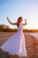 Wall Mural - beautiful slender young woman of Caucasian ethnicity with long hair stands in a white dress in the wind near the sea at sunset