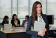 © Bangkok Click Studio - Portrait of young attractive Asian female office worker in formal business suits  smiling at camera in office with blurry colleagues sitting in office as background