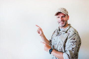 Wall Mural - Handsome man in camouflaged army uniform smiling and looking at the camera pointing with two hands and fingers to the side.