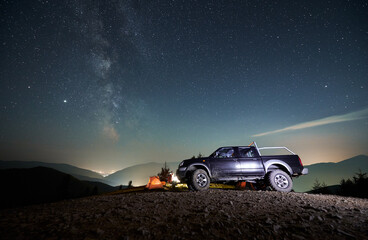 Wall Mural - Black SUV car parked on dirt mountain road near tourist camp with orange tent under night starry sky, against the backdrop of silhouettes of mountain peaks and city lights in the distance.
