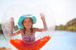 © Ольга Симонова - A girl in a hat stands on the river bank with a transparent inflatable circle in the shape of a heart with orange feathers inside. Beach holidays, swimming, tanning, sunscreens.