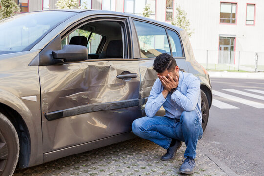 Portrait of man wearing jeans and blue shirt broken his car, being upset and covering his face with palms, dents and scratches on the door of auto, damaged vehicle after accident. Outdoor shot.