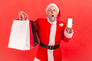 Wall Mural - surprised elderly man wearing santa claus costume holding shopping bags and phone with empty display