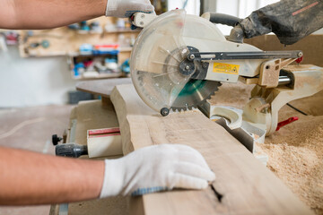 Close up of carpenter cutting with radial arm saw. Woodworker using electric jigsaw in workshop for production of wooden furniture. Joinery work concept.