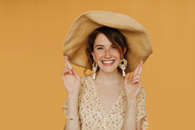 Happy Girl In Flower Cool Dress Crossing Her Fingers On Isolated Backdrop. Positive Lady In Hat And Earrings Smiling On Yellow Background..