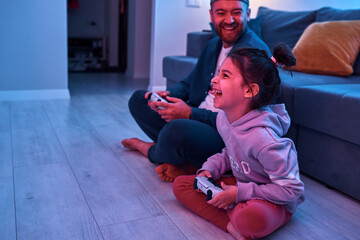 Side view of a joyful daughterwith father playing video game together at home. Dad plays computer games during quarantine with daughter while sitting on the floor in the living room.