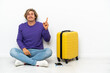 © luismolinero - Young blonde man with suitcase sitting on the floor pointing with the index finger a great idea