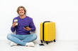 © luismolinero - Young blonde man with suitcase sitting on the floor holding coffee to take away and a mobile