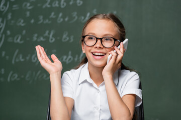 joyful schoolgirl talking on smartphone near blurred chalkboard