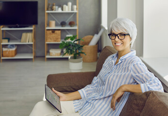 Happy mature woman in glasses enjoying free time on comfortable sofa, holding modern tablet, looking at camera, planning daily task agenda, setting date reminders. Portrait, copy space home background