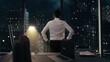 © Iokanan Pro - Back view of thoughtful Businessman standing in his office working late at night with hands on waist, looking out of the window as it rains contemplating his next big idea.