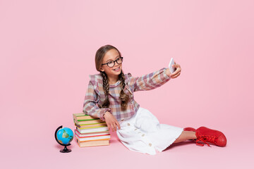 Wall Mural - smiling girl taking selfie while sitting near stack of books and globe on pink background