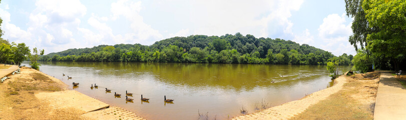 a stunning panoramic shot of the silky brown water of the Chattahoochee river surrounded by lush green trees reflecting off the water with blue sky, clouds and a rainbow at Azalea Park in Roswell GA