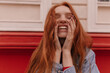 Close-up portrait of young joyful girl having fun outdoors. Pretty lady with long red hair and blue shirt, laughing against bright background
